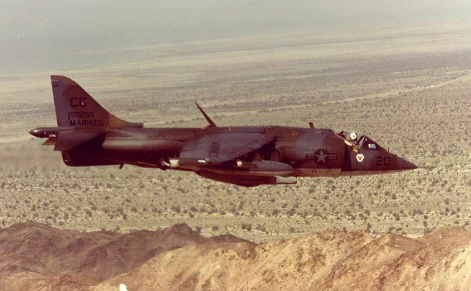 A Harrier jet flies low over a desert landscape with mountains in the foreground.