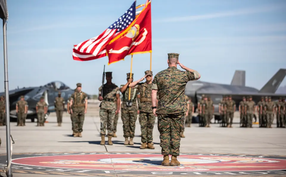 A Marine salutes as others hold the US and Marine Corps flags during a ceremony on an airfield.