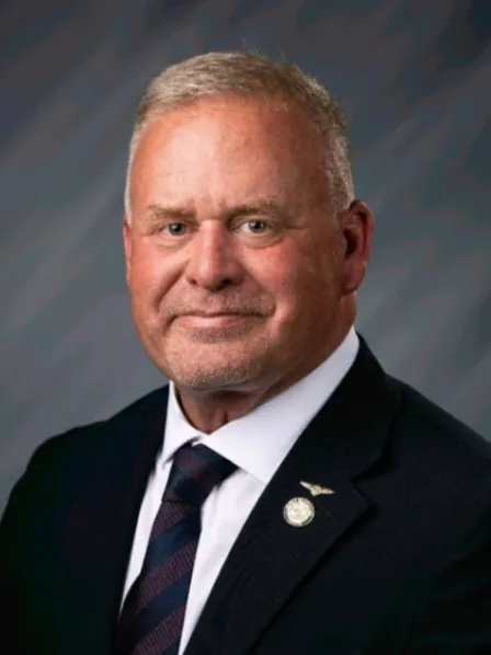 Man in a suit and tie with short gray hair, smiling, in front of a gray background.