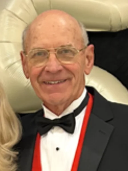 Duane Thiessen in a tuxedo with glasses, standing in front of a large light-colored decoration.