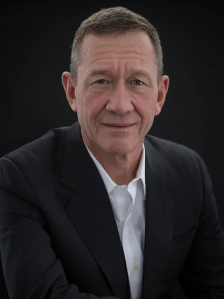 Man in dark suit and white shirt smiling slightly, sitting against a plain black background.