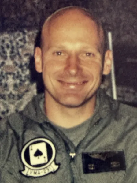 Smiling person in a gray jacket with a patch featuring an ace of spades and the text "IMA-331.