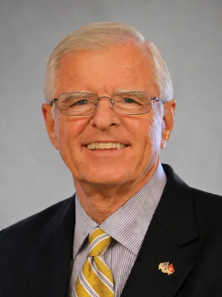 Smiling older man with glasses, gray hair, suit, striped tie, and flag pin on jacket, against gray background.