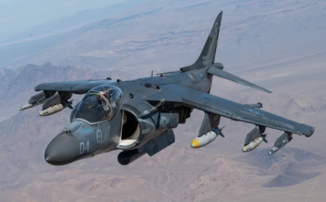A Harrier jet flies over a desert landscape, carrying missiles under its wings.