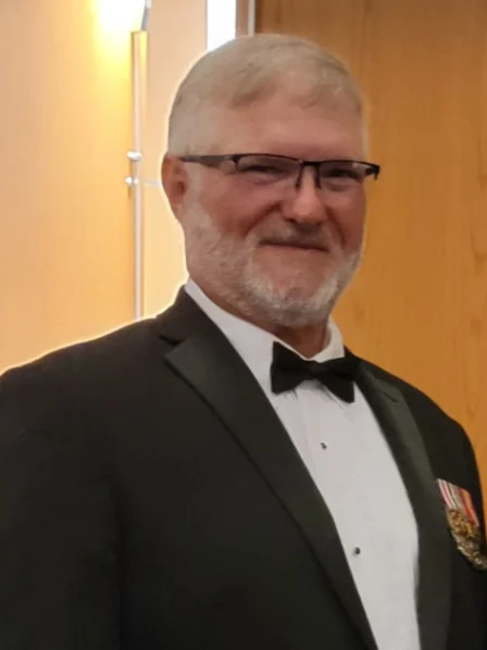 Wally Simmons in a black tuxedo and bow tie, wearing glasses and medals, smiling in a formal setting.
